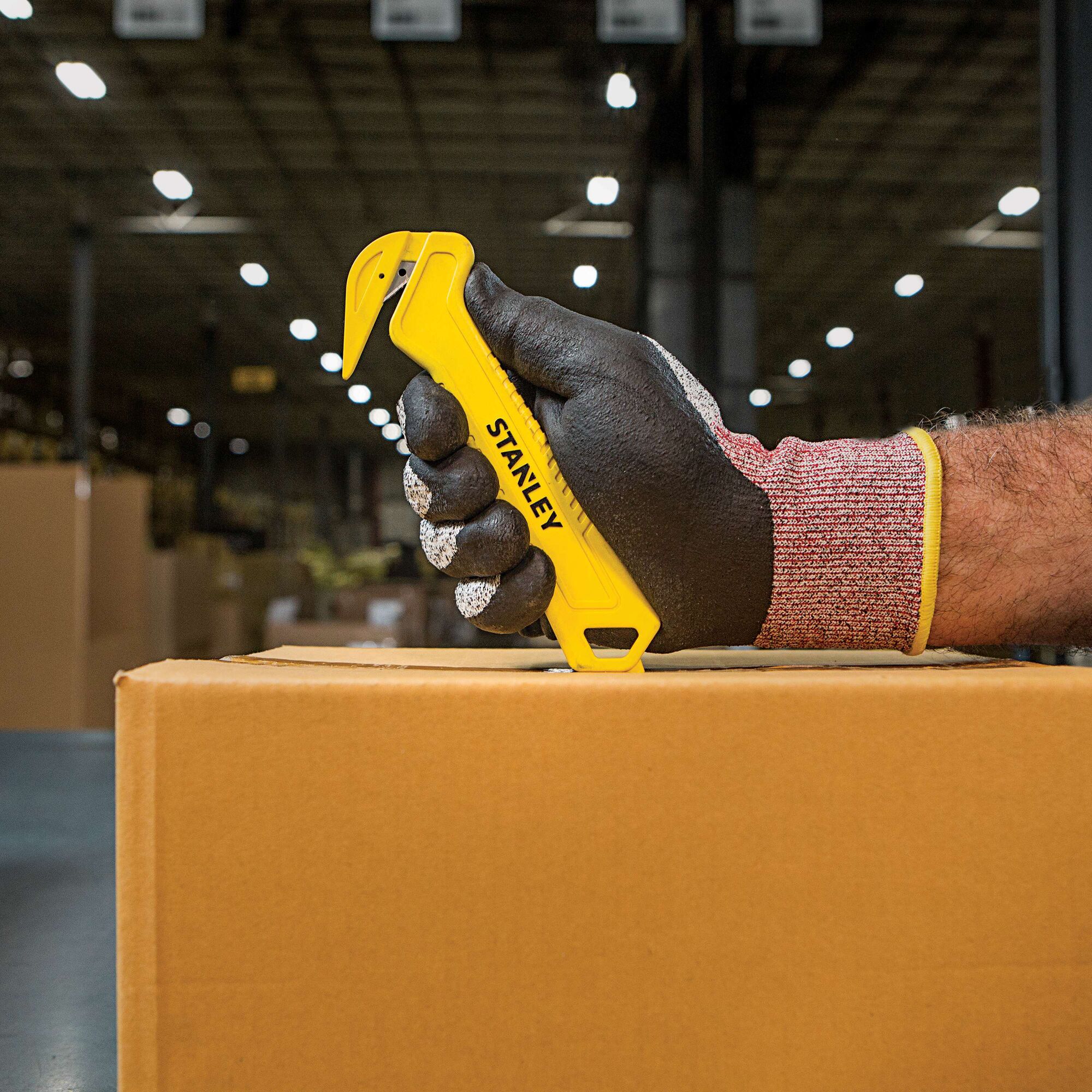 Stanley STHT10355 yellow safety cutter held by a gloved hand slicing a cardboard box in a warehouse.