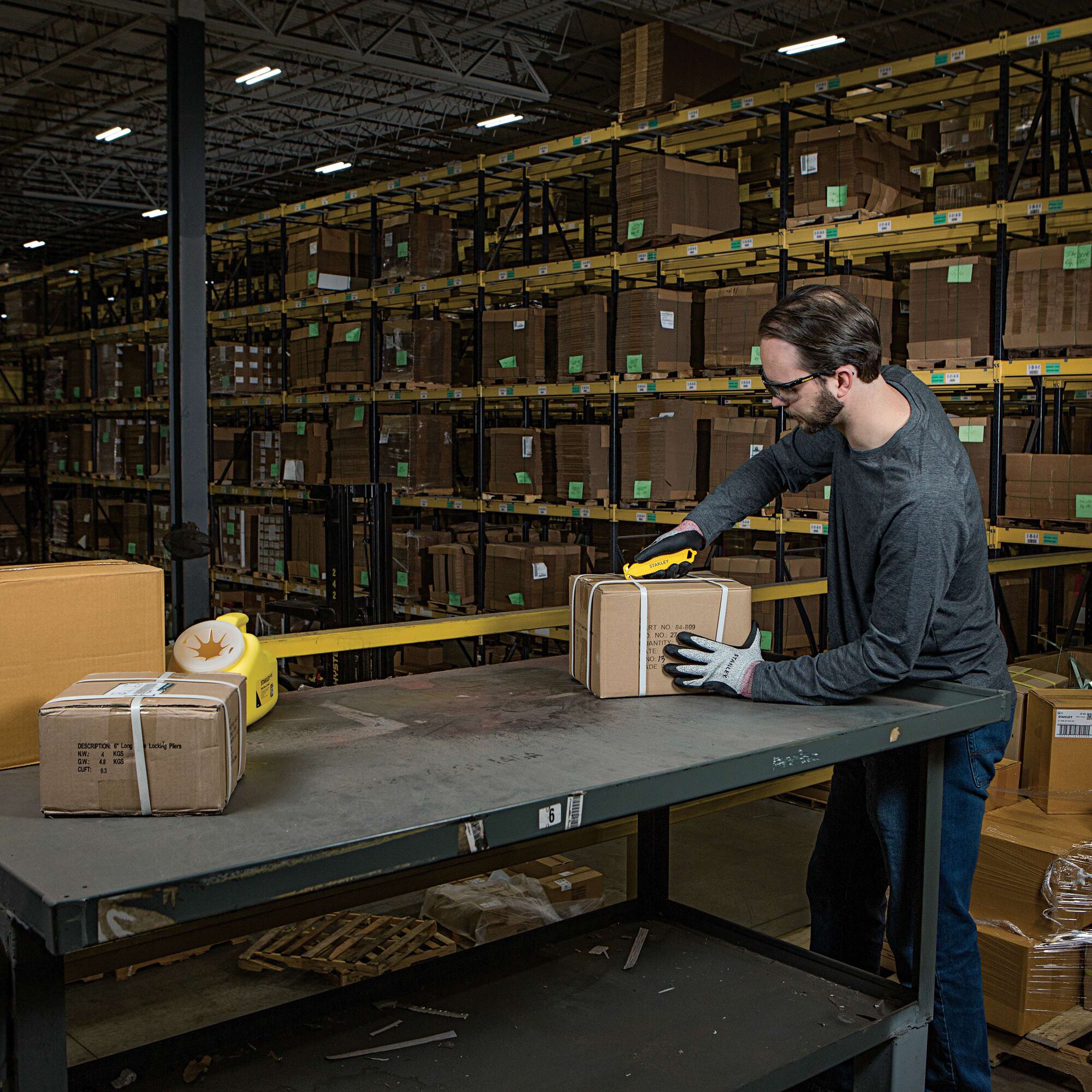 STANLEY STHT10359 utility knife cutting box on warehouse table with shelves of packages in the background.